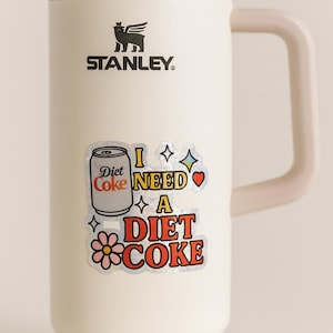 May include: Cream-colored Stanley 90 oz. insulated mug with a handle and a silver lid. The mug features a sticker that says "I NEED A DIET COKE" with a Diet Coke can graphic and other decorative elements. The Stanley logo is visible.