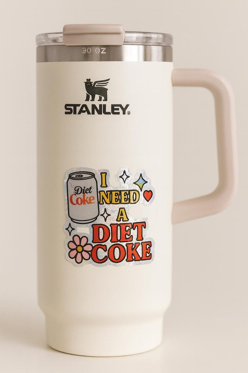 May include: Cream-colored Stanley 90 oz. insulated mug with a handle and a silver lid. The mug features a sticker that says "I NEED A DIET COKE" with a Diet Coke can graphic and other decorative elements. The Stanley logo is visible.