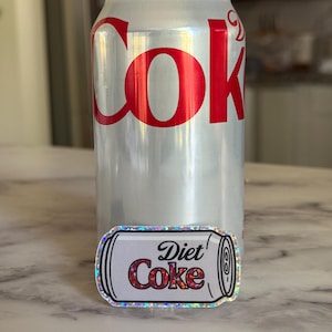 May include: A silver Diet Coke can with red lettering and a Diet Coke sticker. The sticker is a miniature version of the can, with the words "Diet Coke" in a similar font and color scheme.