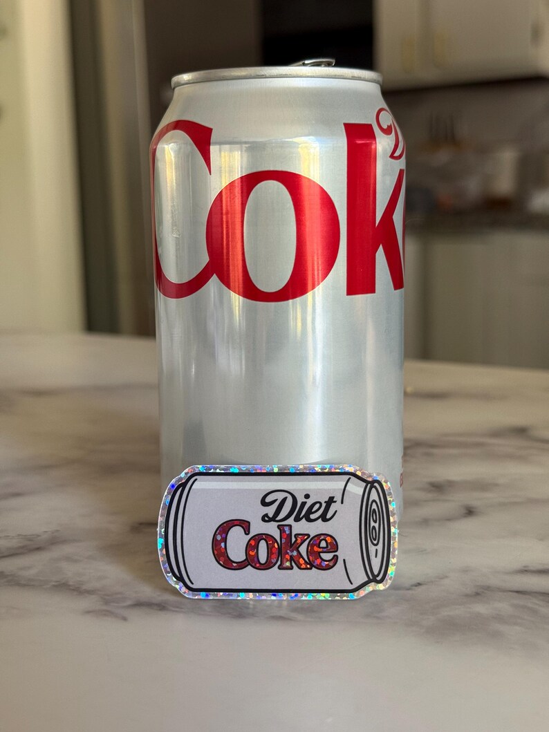 May include: A silver Diet Coke can with red lettering and a Diet Coke sticker. The sticker is a miniature version of the can, with the words "Diet Coke" in a similar font and color scheme.