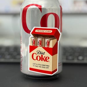 May include: A silver Diet Coke can with red lettering and a sticker of a cigarette box. The sticker reads "Diet Coke" and "Fridge Cigs" with the tagline "Just a little something to take the edge off."