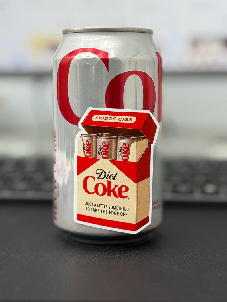 May include: A silver Diet Coke can with red lettering and a sticker of a cigarette box. The sticker reads "Diet Coke" and "Fridge Cigs" with the tagline "Just a little something to take the edge off."