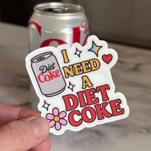 May include: A Diet Coke sticker with the phrase "I NEED A DIET COKE" in red, yellow, and white lettering. The sticker features a cartoon image of a Diet Coke can and a pink flower. A silver Diet Coke can is in the background.