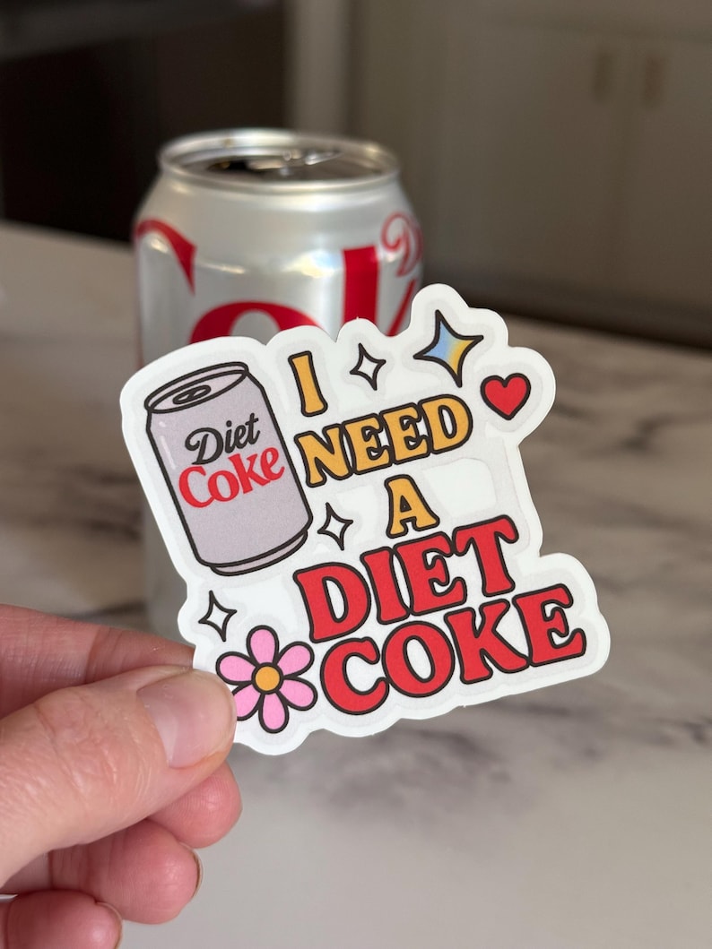 May include: A Diet Coke sticker with the phrase "I NEED A DIET COKE" in red, yellow, and white lettering. The sticker features a cartoon image of a Diet Coke can and a pink flower. A silver Diet Coke can is in the background.