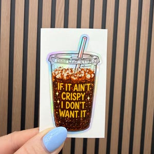 May include: A holographic sticker featuring a clear plastic cup filled with dark brown iced coffee and ice cubes. A straw sticks out of the lid. The text IF IT AIN'T CRISPY I DON'T WANT IT is printed in yellow on the front of the cup.