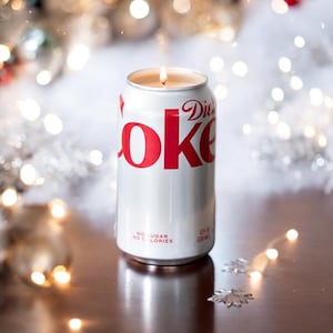 Può includere: Una candela bianca a forma di lattina di Coca-Cola Light con un logo "Coke" rosso e le parole "No Sugar No Calories" sul fondo. La candela è accesa e la fiamma è visibile.