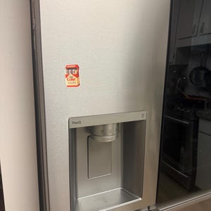 K&ouml;nnte beinhalten: Edelstahlk&uuml;hlschrank mit Wasser- und Eisspender. Auf der T&uuml;r befindet sich ein kleiner Coca-Cola-Aufkleber. Der K&uuml;hlschrank hat ein schlankes, modernes Design mit einer schwarzen Glast&uuml;r auf der rechten Seite.