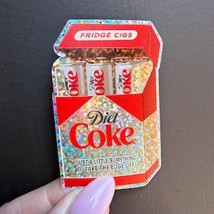 May include: A holographic sticker shaped like a red cigarette pack with the text FRIDGE CIGS at the top. Inside are three small cans labeled Diet Coke. The front features the text Diet Coke and JUST A LITTLE SOMETHING TO TAKE THE EDGE OFF.
