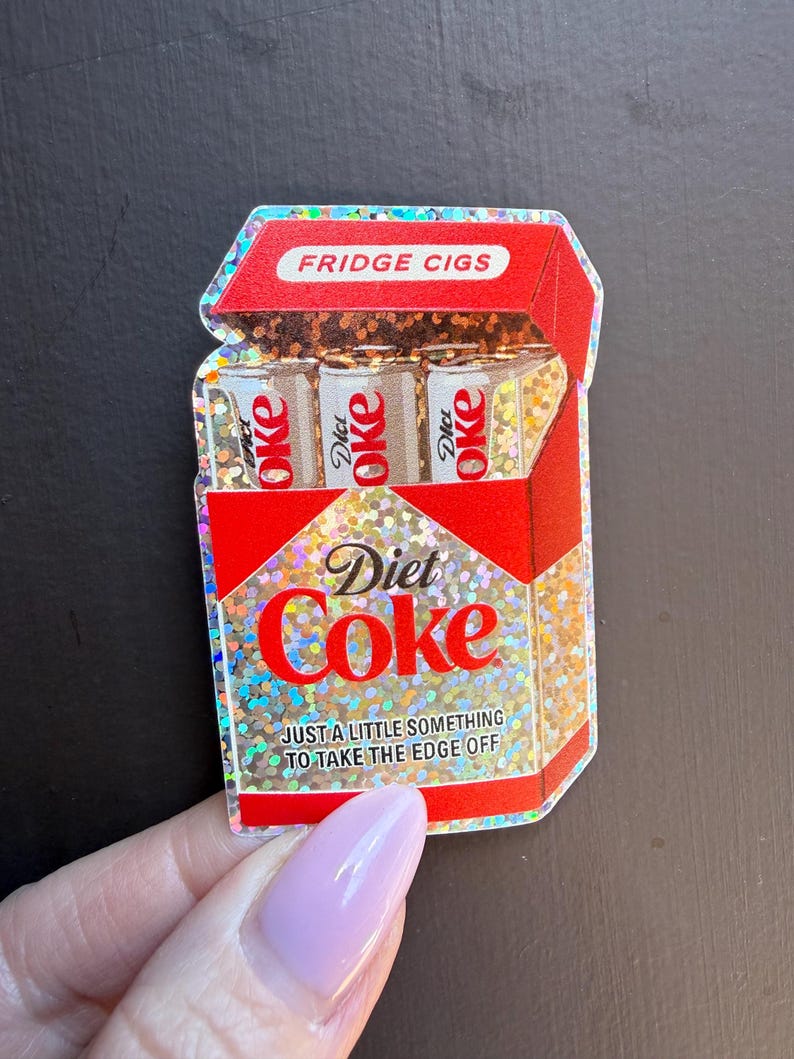 May include: A holographic sticker shaped like a red cigarette pack with the text FRIDGE CIGS at the top. Inside are three small cans labeled Diet Coke. The front features the text Diet Coke and JUST A LITTLE SOMETHING TO TAKE THE EDGE OFF.