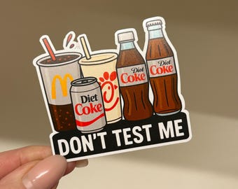 May include: A die-cut sticker featuring a McDonald's soda cup, a Chick-fil-A cup, a Diet Coke can, and two Diet Coke glass bottles. The text DON'T TEST ME is printed in white on a black rectangular banner at the bottom of the sticker design.