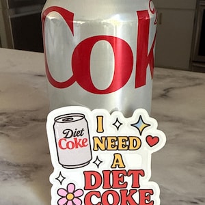 May include: A silver Diet Coke can with red lettering stands behind a sticker that says "I NEED A DIET COKE." The sticker features a Diet Coke can graphic, stars, a heart, and a flower.