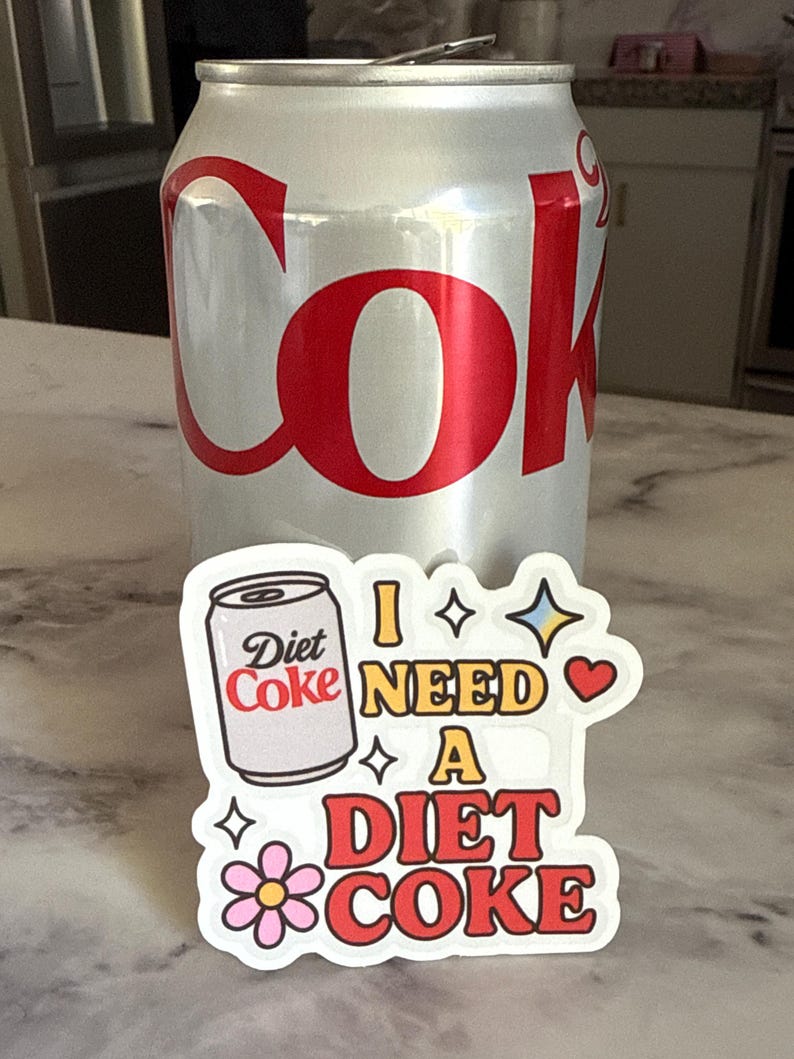 May include: A silver Diet Coke can with red lettering stands behind a sticker that says "I NEED A DIET COKE." The sticker features a Diet Coke can graphic, stars, a heart, and a flower.