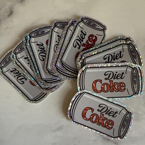 May include: A collection of Diet Coke stickers. The stickers are shaped like silver soda cans with the words "Diet Coke" in red and white. The stickers have a holographic, iridescent border.