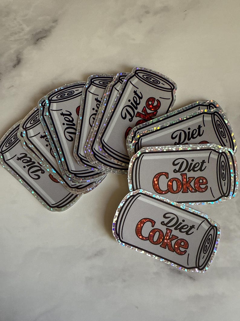 May include: A collection of Diet Coke stickers. The stickers are shaped like silver soda cans with the words "Diet Coke" in red and white. The stickers have a holographic, iridescent border.