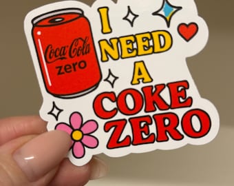 May include: A die-cut sticker featuring a red Coca-Cola Zero can illustration next to the yellow and red text I NEED A COKE ZERO. The design includes a pink flower, a red heart, and two small star icons on a white background.
