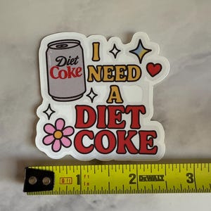 May include: A white sticker with a Diet Coke can illustration and the phrase "I NEED A DIET COKE" in red and yellow. The sticker is approximately 2 inches wide, with a flower, stars, and a heart design.