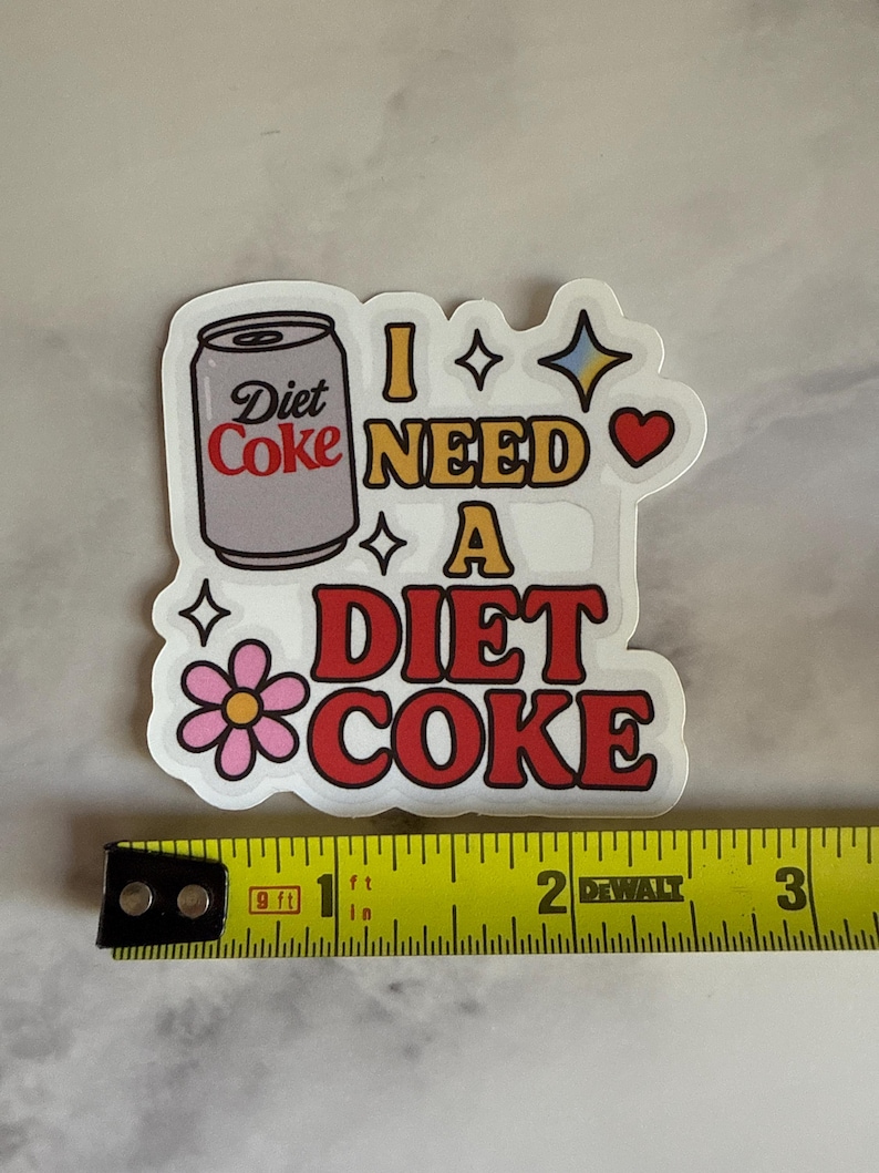 May include: A white sticker with a Diet Coke can illustration and the phrase "I NEED A DIET COKE" in red and yellow. The sticker is approximately 2 inches wide, with a flower, stars, and a heart design.