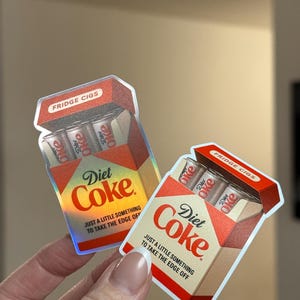 K&ouml;nnte beinhalten: Zwei Aufkleber in Form von Diet Coke Zigarettenpackungen. Die Packungen sind rot, wei&szlig; und beige mit den Aufdrucken "Diet Coke" und "Fridge Cigs". Jede Packung enth&auml;lt drei Diet Coke Dosen. Der Text "Nur ein kleines bisschen, um die Kanten abzurunden" ist ebenfalls aufgedruckt.