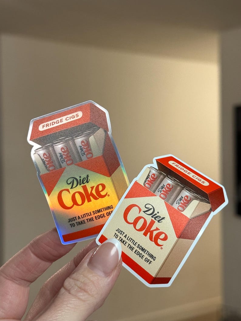 K&ouml;nnte beinhalten: Zwei Aufkleber in Form von Diet Coke Zigarettenpackungen. Die Packungen sind rot, wei&szlig; und beige mit den Aufdrucken "Diet Coke" und "Fridge Cigs". Jede Packung enth&auml;lt drei Diet Coke Dosen. Der Text "Nur ein kleines bisschen, um die Kanten abzurunden" ist ebenfalls aufgedruckt.