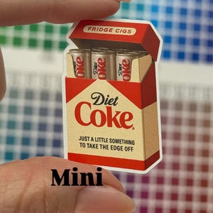 K&ouml;nnte beinhalten: Ein Aufkleber, der eine Packung "Fridge Cigs" mit drei "Diet Coke" Dosen zeigt. Die Verpackung ist rot und beige mit den Worten "Diet Coke" und "Just a little something to take the edge off". Das Wort "Mini" steht unten.