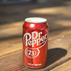May include: A red aluminum can of Dr Pepper soda with the text "Dr Pepper Est. 1885" and "23 Authentic Blend of 23 Flavors" printed on the can. The can is 12 fluid ounces (355 ml).
