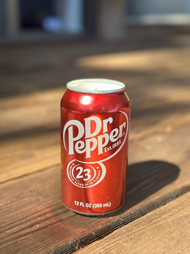 May include: A red aluminum can of Dr Pepper soda with the text "Dr Pepper Est. 1885" and "23 Authentic Blend of 23 Flavors" printed on the can. The can is 12 fluid ounces (355 ml).
