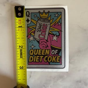 May include: A sticker featuring a playing card design with a Diet Coke can held by a hand. The text "Queen of Diet Coke" is displayed in a retro font. The sticker measures approximately 3 inches in height.
