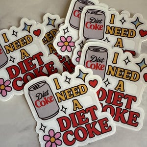 May include: A collection of stickers with a retro design. Each sticker features a Diet Coke can illustration and the phrase "I NEED A DIET COKE" in red and white lettering. Decorative elements include stars, hearts, and flowers.