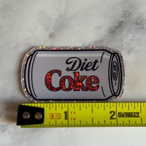 May include: A Diet Coke sticker with a silver outline. The text "Diet Coke" is in red and black. The sticker is approximately 2 inches long.