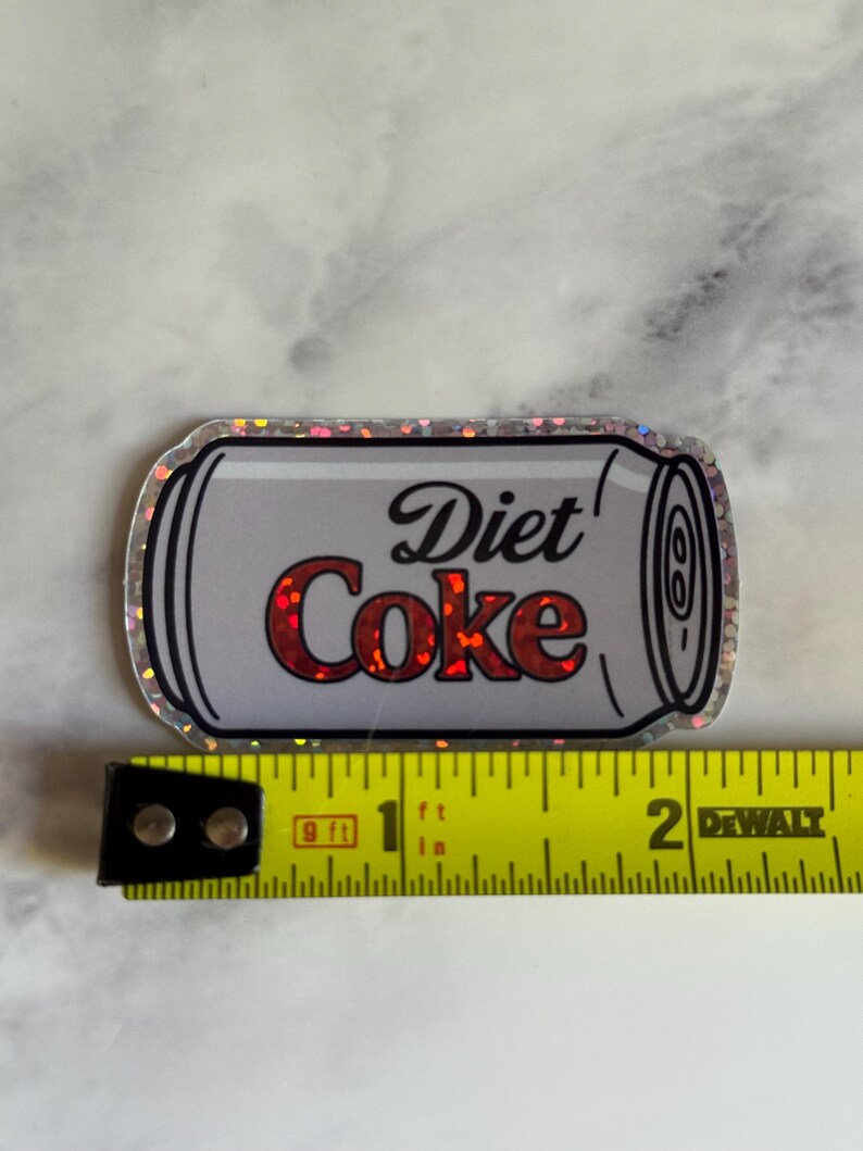 May include: A Diet Coke sticker with a silver outline. The text "Diet Coke" is in red and black. The sticker is approximately 2 inches long.