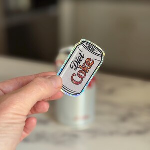 May include: A Diet Coke sticker in the shape of a soda can. The sticker has a white background with the words "Diet Coke" in a stylized font. The letters are filled with a floral pattern. The sticker has a holographic border.