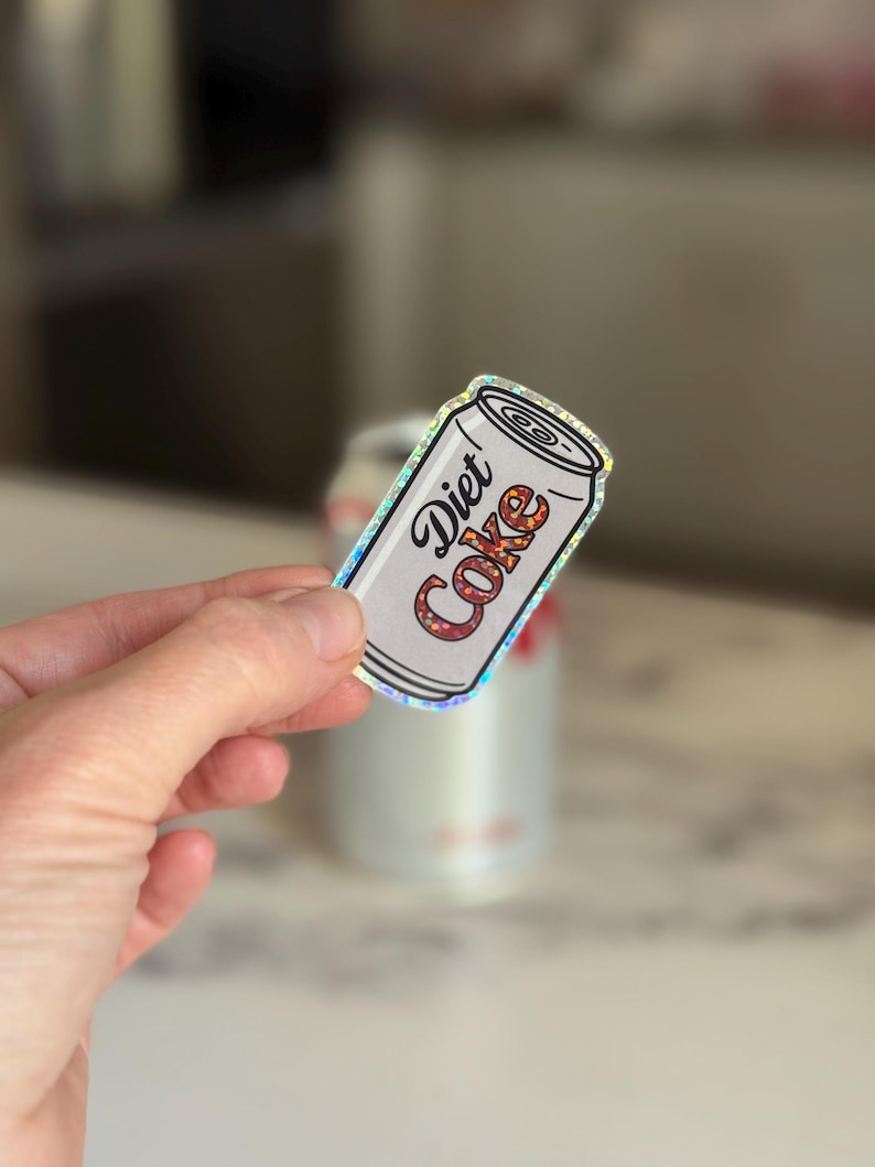 May include: A Diet Coke sticker in the shape of a soda can. The sticker has a white background with the words "Diet Coke" in a stylized font. The letters are filled with a floral pattern. The sticker has a holographic border.