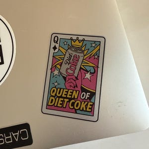 May include: A sticker featuring a playing card design with the text "Queen of Diet Coke." The card shows a Diet Coke can held by a hand, with a crown and starburst design. The card has a pink, blue, and yellow color scheme.