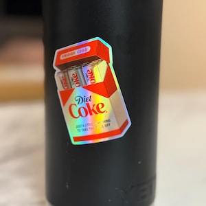 K&ouml;nnte beinhalten: Ein holografischer Aufkleber einer Diet Coke Zigarettenschachtel mit dem Text "Fridge Cigs" und "Diet Coke". Der Aufkleber hat auch den Text "Just a little something to take the edge off". Der Aufkleber befindet sich auf einem schwarzen Becher.