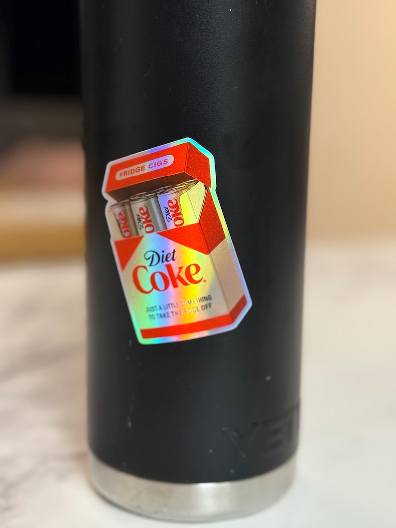 K&ouml;nnte beinhalten: Ein holografischer Aufkleber einer Diet Coke Zigarettenschachtel mit dem Text "Fridge Cigs" und "Diet Coke". Der Aufkleber hat auch den Text "Just a little something to take the edge off". Der Aufkleber befindet sich auf einem schwarzen Becher.