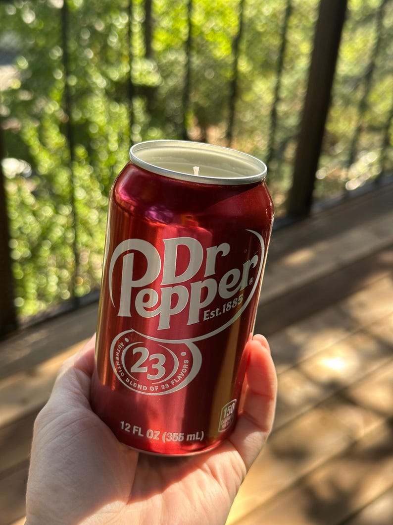 May include: A red aluminum can candle with a Dr Pepper label. The label features the Dr Pepper logo, the text "Est. 1885", and a circle with the number "23" and the text "Authentic Blend of 23 Flavors". The can is 12 fluid ounces (355 ml).