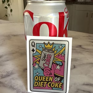 May include: A silver Diet Coke can stands behind a playing card-style sticker. The sticker features a Diet Coke can with a crown, the words "Queen of Diet Coke," and a colorful design with pink, blue, and yellow accents.