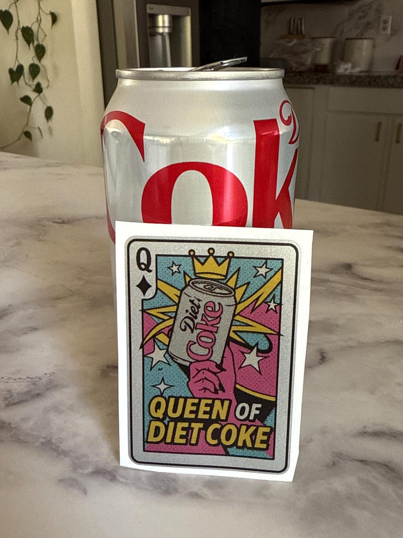 May include: A silver Diet Coke can stands behind a playing card-style sticker. The sticker features a Diet Coke can with a crown, the words "Queen of Diet Coke," and a colorful design with pink, blue, and yellow accents.