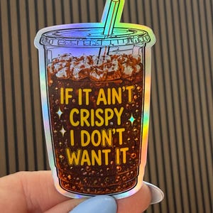 May include: A holographic sticker shaped like a plastic cup filled with a dark brown carbonated beverage, ice cubes, and a straw. The text IF IT AIN'T CRISPY I DON'T WANT IT is printed in yellow letters on the front of the cup with small star accents.