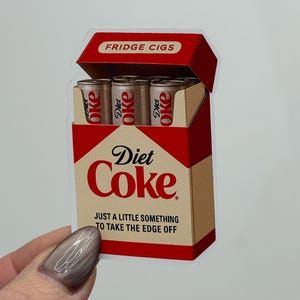 K&ouml;nnte beinhalten: Ein Aufkleber, der eine Diet Coke-Zigarettenschachtel darstellt. Die rote und beige Packung tr&auml;gt die Aufschrift "FRIDGE CIGS" und "Diet Coke". Im Inneren befinden sich drei Miniatur-Diet Coke-Dosen. Der Text "JUST A LITTLE SOMETHING TO TAKE THE EDGE OFF" ist ebenfalls sichtbar.