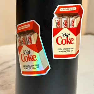 K&ouml;nnte beinhalten: Zwei Aufkleber mit einem Retro-Design, das einer Zigarettenschachtel &auml;hnelt, mit der Aufschrift "Fridge Cigs" und "Diet Coke". Die Aufkleber zeigen eine rot-wei&szlig;e Verpackung mit dem Markennamen "Diet Coke" und dem Satz "Nur ein kleines bisschen, um die Kanten abzurunden."