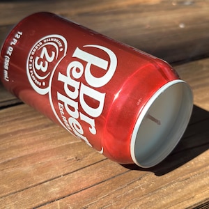 May include: A red aluminum can candle with a white label that says "Dr Pepper" and "23 Flavors" with a white wick sticking out of the top.