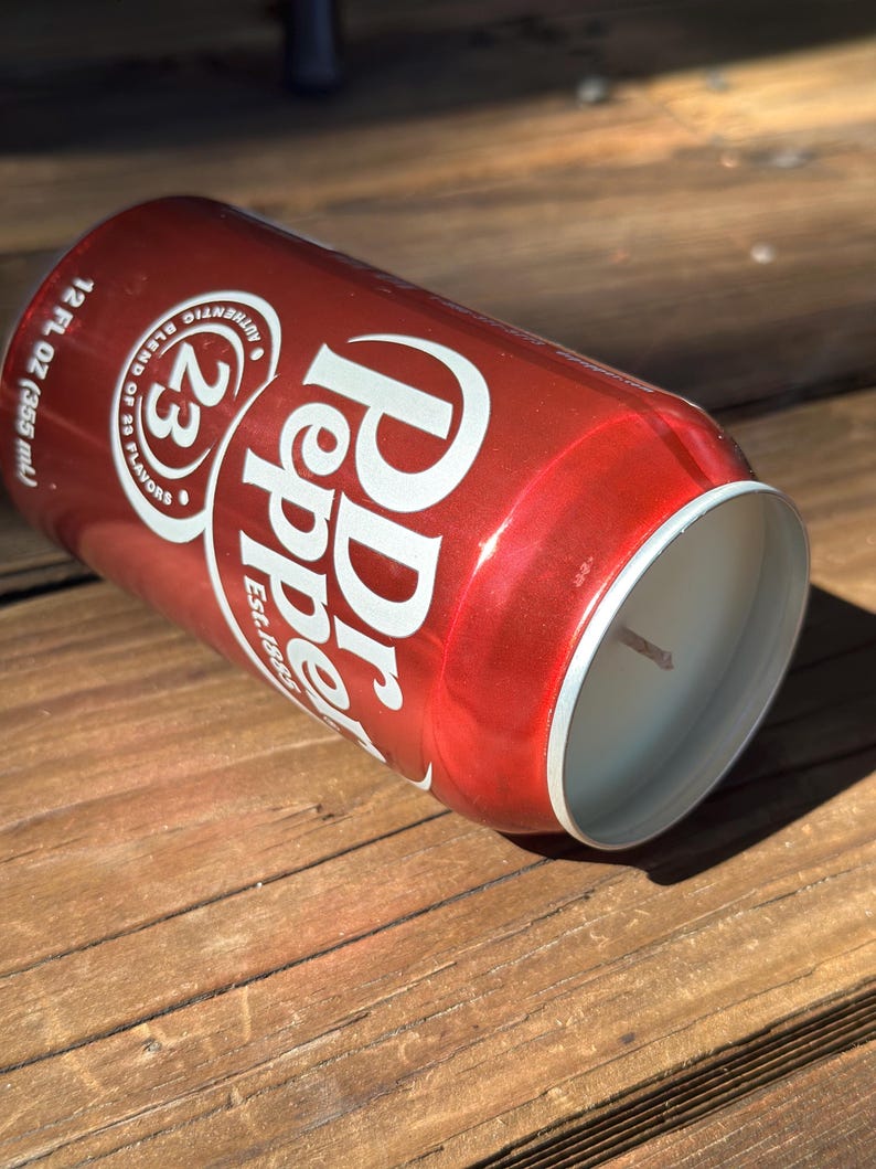 May include: A red aluminum can candle with a white label that says "Dr Pepper" and "23 Flavors" with a white wick sticking out of the top.