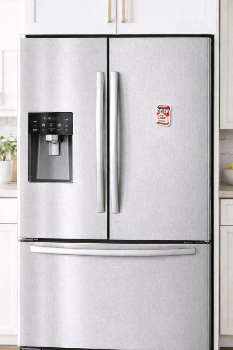 K&ouml;nnte beinhalten: Ein K&uuml;hlschrank aus Edelstahl mit einem rot-wei&szlig;en Coke-Magneten an der T&uuml;r. Der K&uuml;hlschrank hat einen Wasser- und Eisspender und eine untere Gefrierschublade. Die Griffe sind silberfarben.
