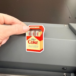 May include: A Diet Coke sticker in the shape of a cigarette pack. The sticker is red, yellow, and white, with the words "Diet Coke" and "Just a little something to take the edge off." The top of the pack reads "Fridge Cigs."