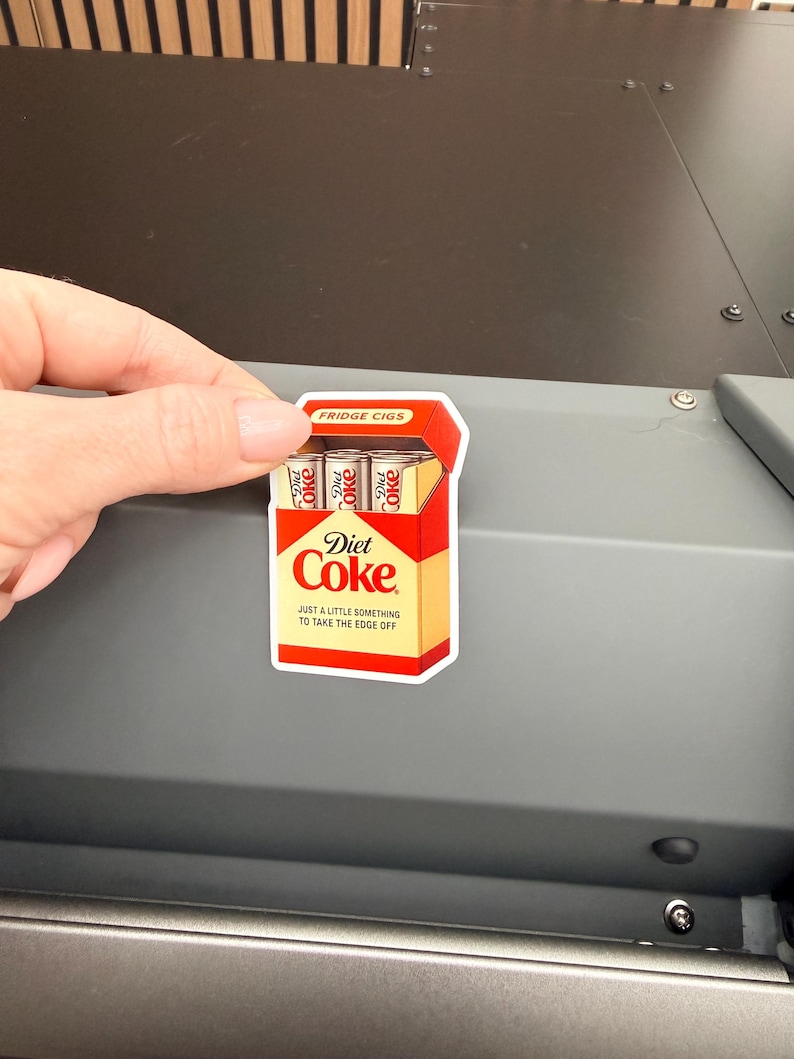 May include: A Diet Coke sticker in the shape of a cigarette pack. The sticker is red, yellow, and white, with the words "Diet Coke" and "Just a little something to take the edge off." The top of the pack reads "Fridge Cigs."