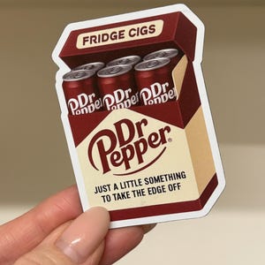 May include: A die-cut sticker shaped like a cigarette pack, colored in maroon and cream. It features the text FRIDGE CIGS at the top, Dr Pepper branding, and the phrase JUST A LITTLE SOMETHING TO TAKE THE EDGE OFF. The pack contains five red soda cans.