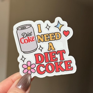 May include: A sticker with a Diet Coke can illustration and the text "I NEED A DIET COKE" in red and yellow. The sticker includes a pink flower, stars, and a heart.
