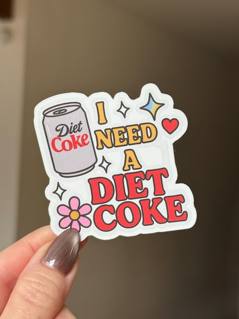 May include: A sticker with a Diet Coke can illustration and the text "I NEED A DIET COKE" in red and yellow. The sticker includes a pink flower, stars, and a heart.