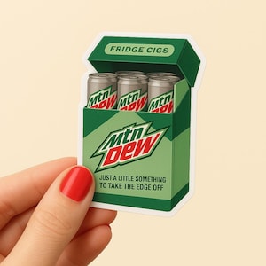 May include: A sticker depicting a green box labeled "FRIDGE CIGS" with three silver cans of "Mtn Dew" inside. The box has the "Mtn Dew" logo and the text "JUST A LITTLE SOMETHING TO TAKE THE EDGE OFF".